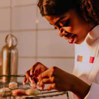 Crie uma uma imagem realista de uma jovem confeiteira negra confeccionando macarons numa cozinha com bancada de mármore e armários brancos 